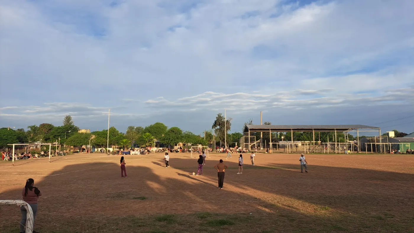 Chicas jugando al fútbol al atardecer en la cancha de tierra del polideportivo, con familias mirando desde el costado